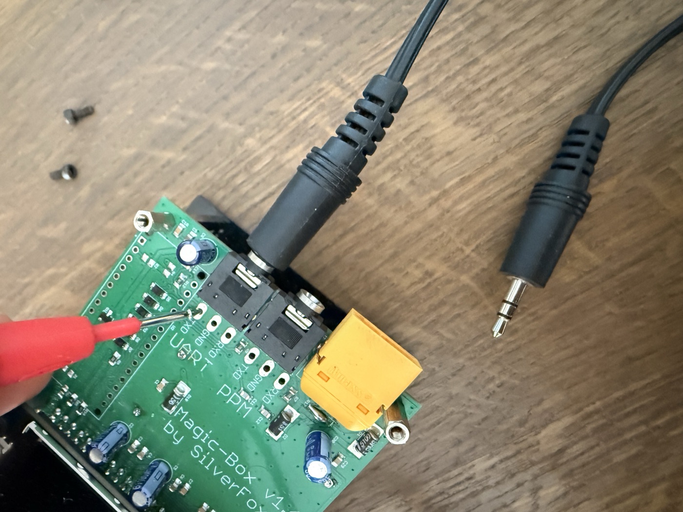 Close-up of the UART pads with a red multimeter probe pointing at the TXD pad, with a 3.5 mm stereo plug nearby for reference